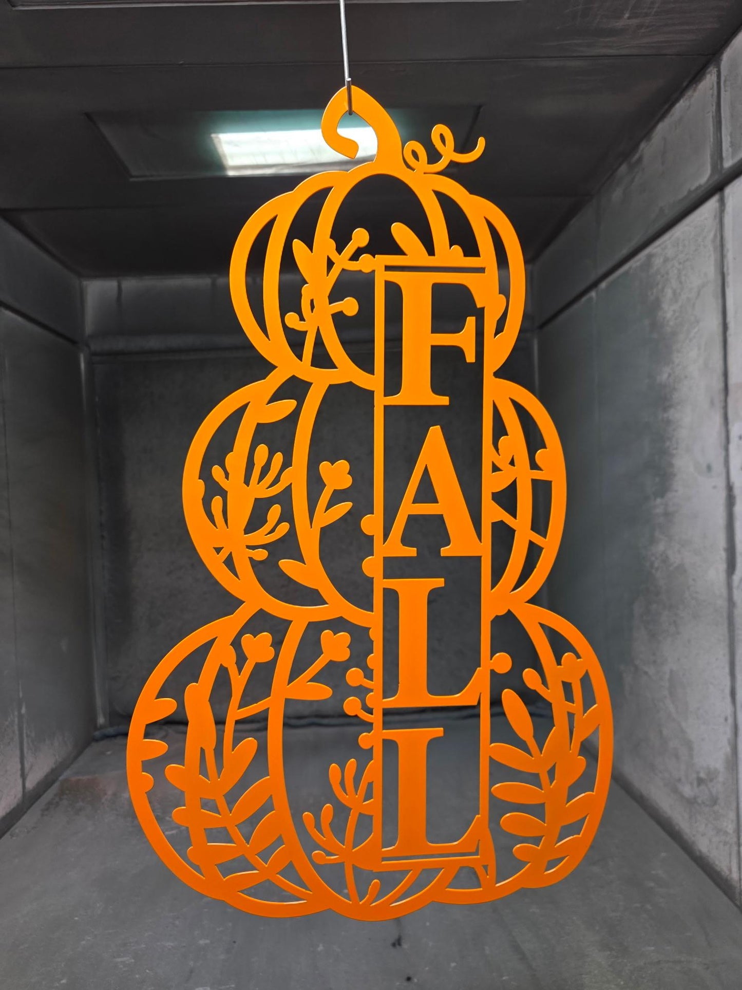 An orange laser-cut metal gourd decoration featuring the word 'FALL' vertically, hanging in a dark industrial storage room.