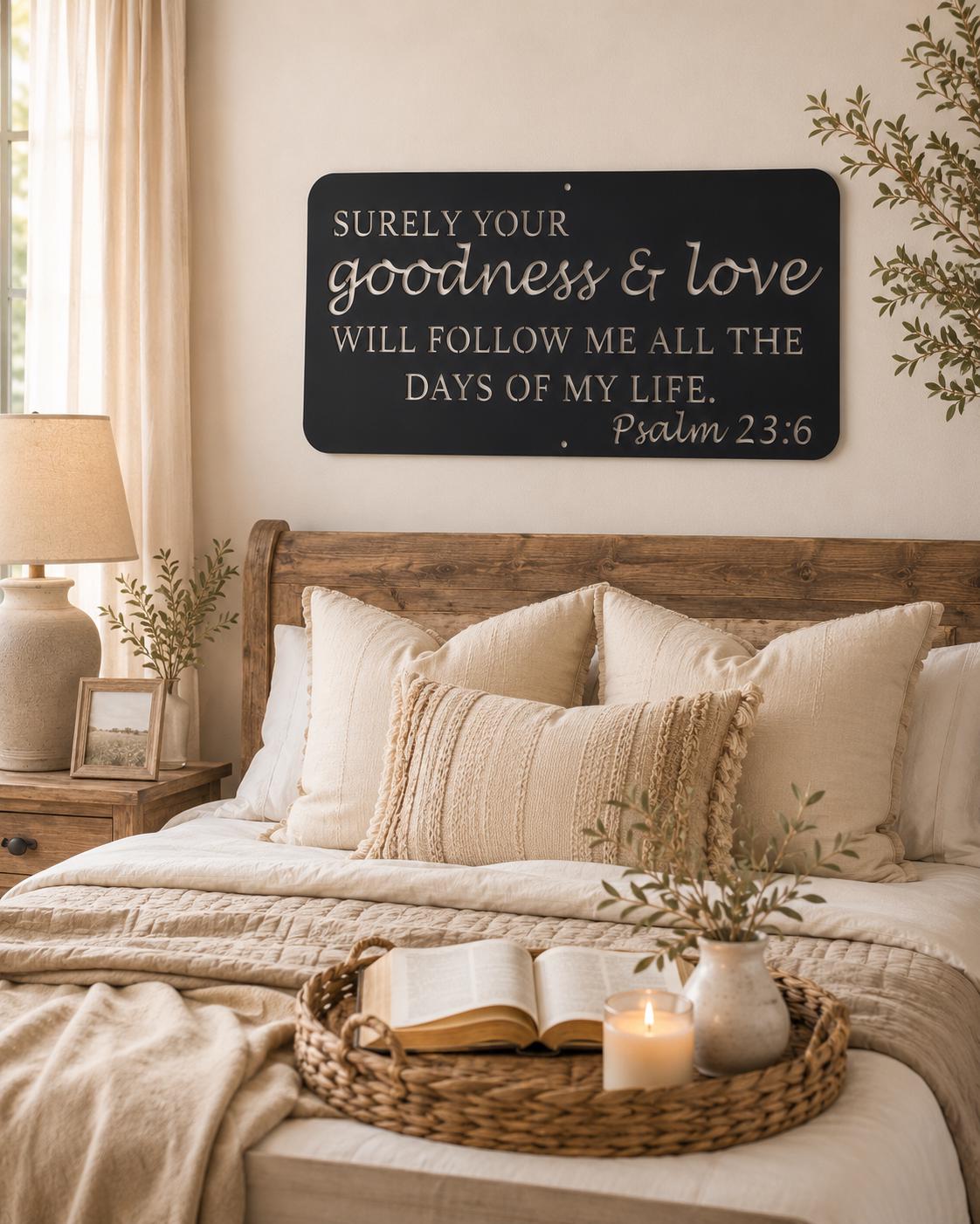 A styled bedroom scene with a wooden bed dressed in neutral beige linens, a woven tray with an open book and candle on the foot of the bed, and a black wall sign above the headboard that reads, 'Surely your goodness & love will follow me all the days of my life. Psalm 23:6'.
