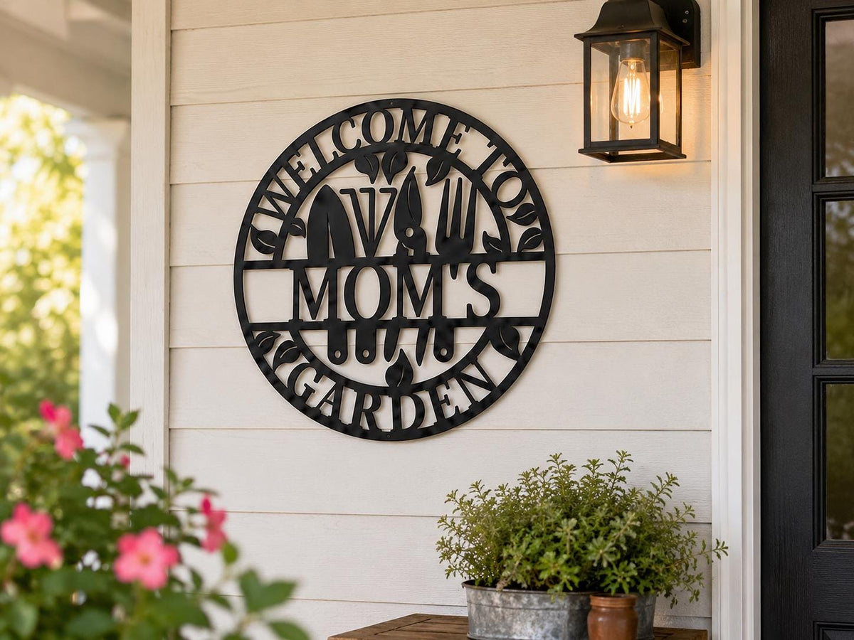 A circular black metal wall sign reading 'Welcome to Mom's Vegarden' with silhouettes of a cow, carrots, and garden tools, mounted on a white shiplap wall on a porch next to a lit lantern and potted plants.