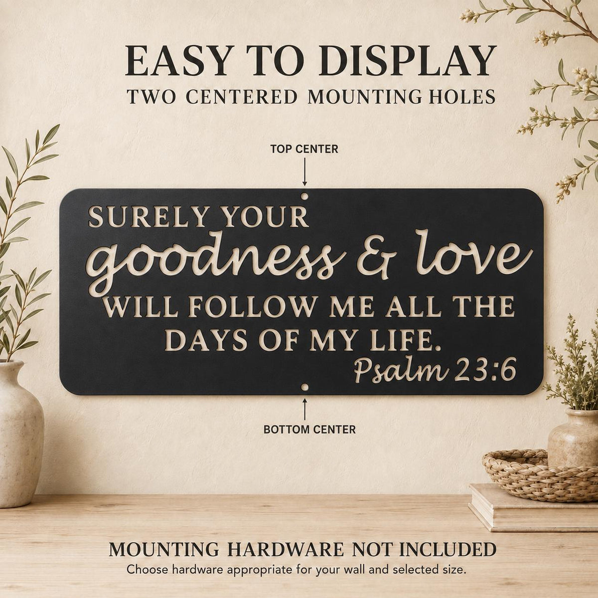 A black rectangular wall sign with cream lettering that reads, 'SURELY YOUR goodness & love WILL FOLLOW ME ALL THE DAYS OF MY LIFE. Psalm 23:6,' shown on a beige wall above a wooden surface with decorative plants and baskets.