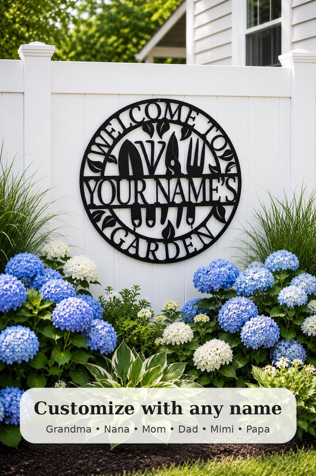 A circular black metal personalized garden sign mounted on a white fence, surrounded by blue and white hydrangeas and green foliage. The sign reads 'WELCOME TO YOUR NAME'S GARDEN' with a silhouette of a garden hose spraying water.