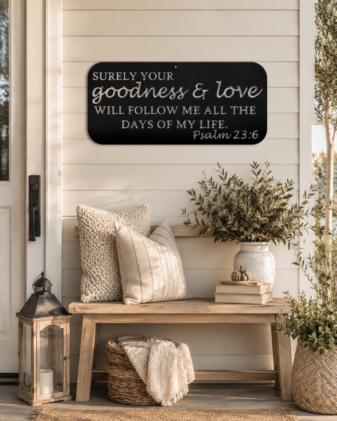 A black rectangular wall sign with white script text reading 'Surely your goodness & love will follow me all the days of my life. Psalm 23:6' displayed on a white shiplap wall above a rustic wooden bench with pillows, books, and a lantern.