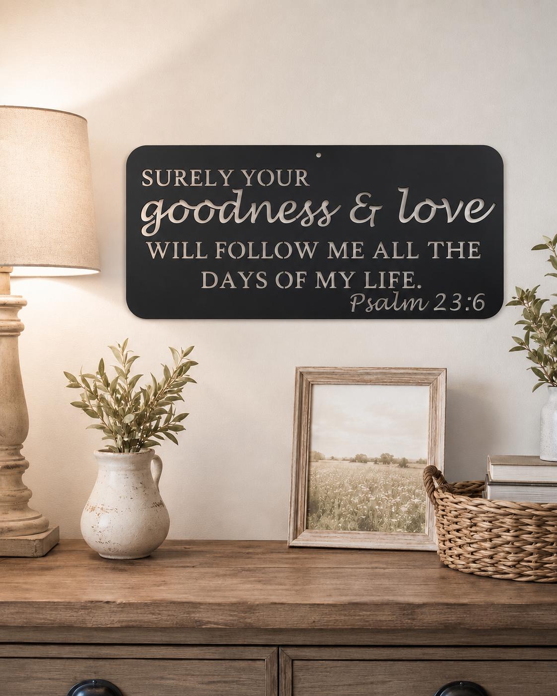 A black rectangular wall sign with white script and serif text reading 'SURELY YOUR goodness & love WILL FOLLOW ME ALL THE DAYS OF MY LIFE. Psalm 23:6' displayed above a wooden console table with a lamp, vase, framed picture, and basket.