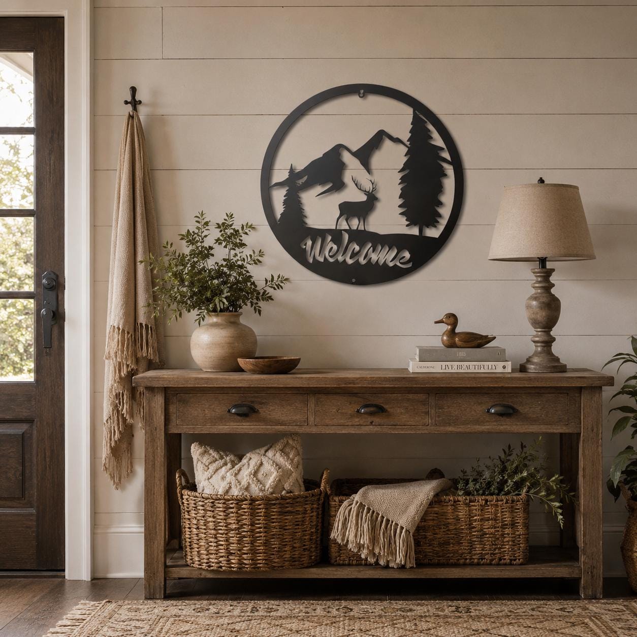 A rustic black metal wall sign featuring a mountain, tree silhouette, and the word 'Welcome' in script, displayed above a wooden console table in a styled home entryway.