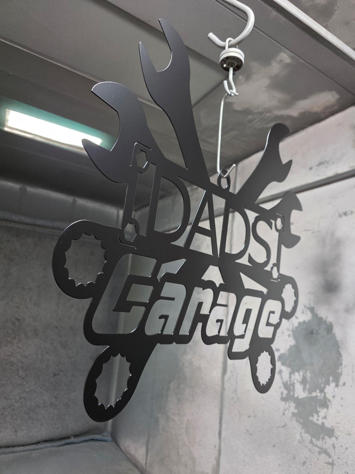 A black metal garage sign reading 'DAD'S Garage' with wrench and gear shapes, hanging from a hook in an industrial-style interior.
