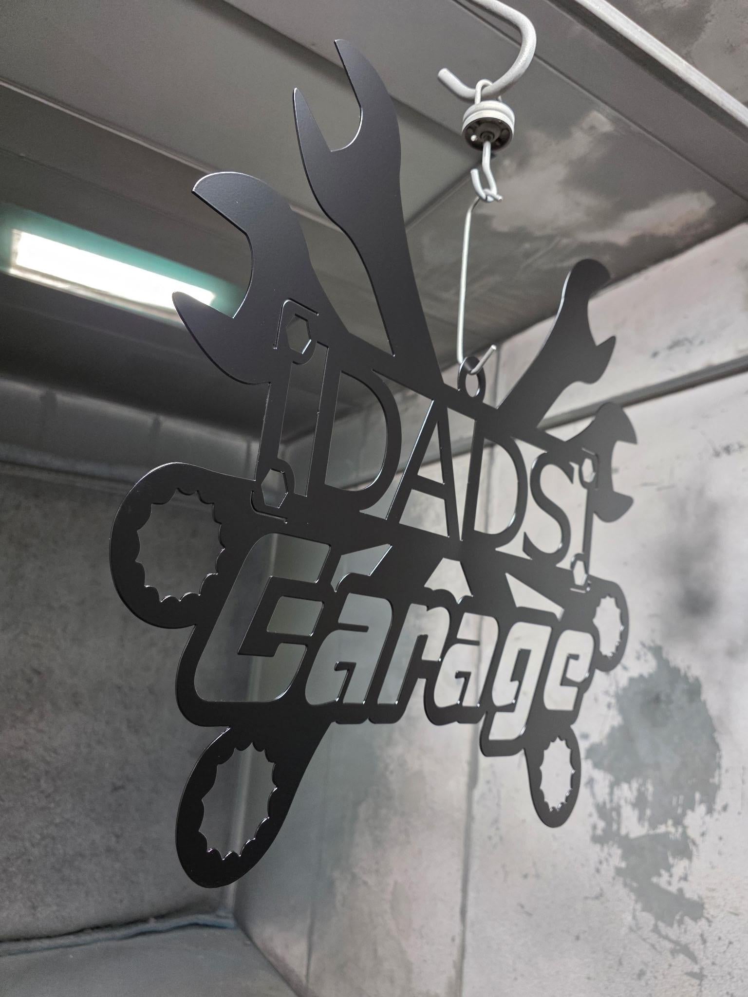 A black metal garage sign reading 'DAD'S Garage' with wrench and gear shapes, hanging from a hook in an industrial-style interior.