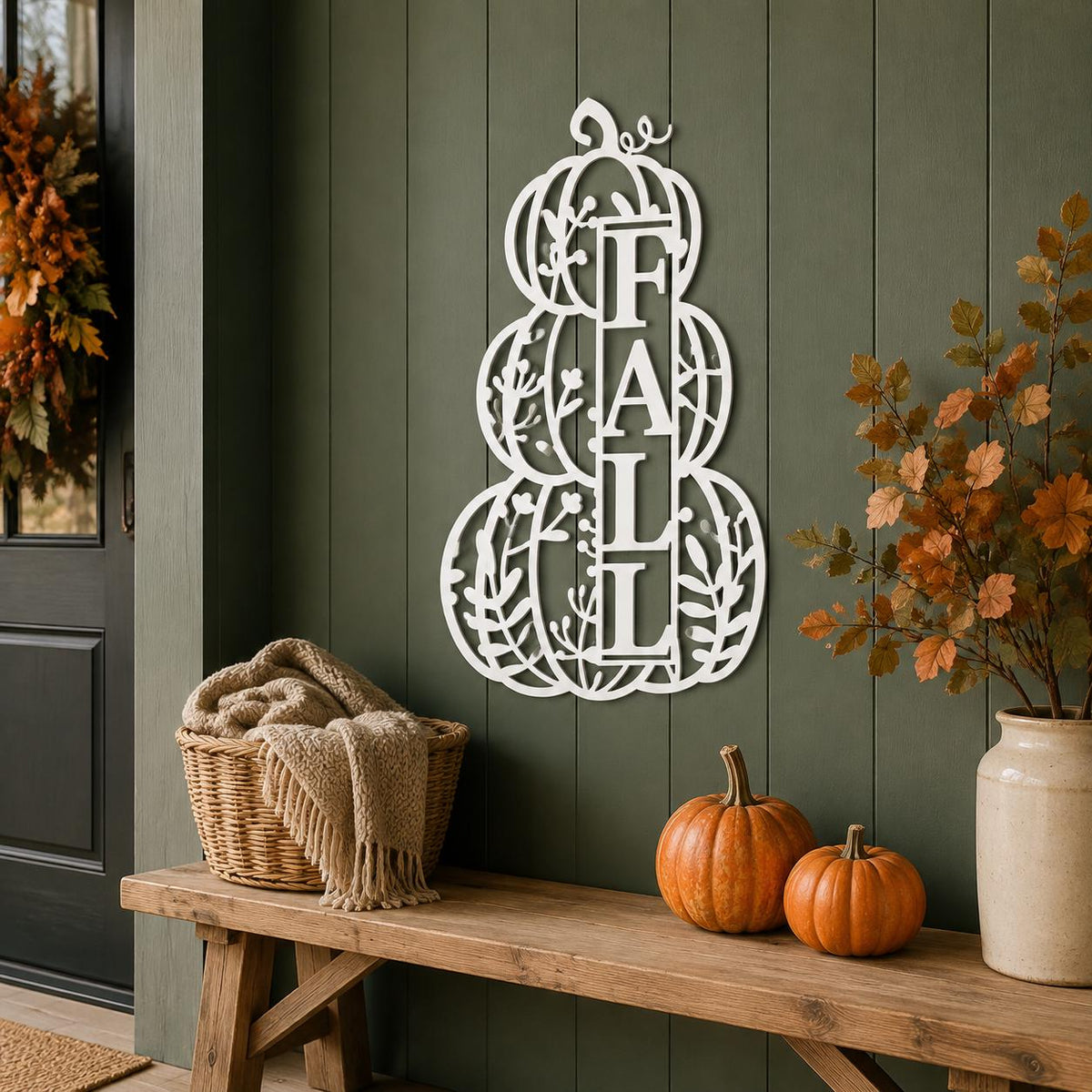 White metal fall wreath wall decor featuring the word 'FALL' integrated into stacked pumpkins and leaves, displayed on a dark green paneled wall above a rustic wooden bench with pumpkins and a basket.