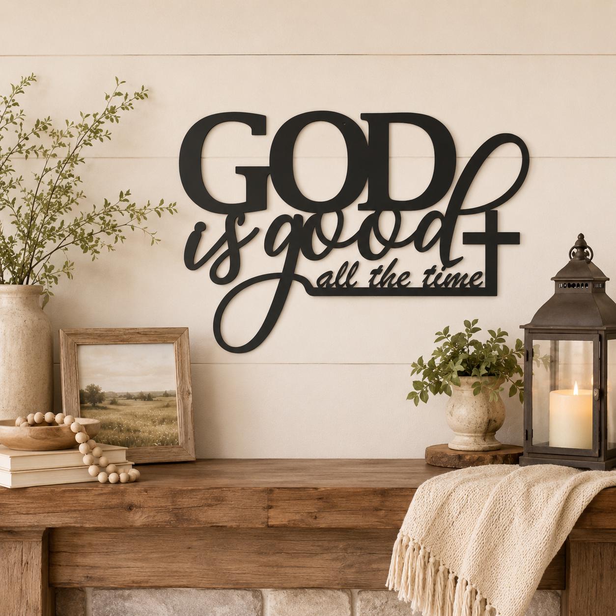 Black metal wall art reading 'GOD is good all the time' with a cross, displayed on a cream shiplap wall above a rustic wooden mantel decorated with a candle lantern, plants, beads, and a throw blanket.