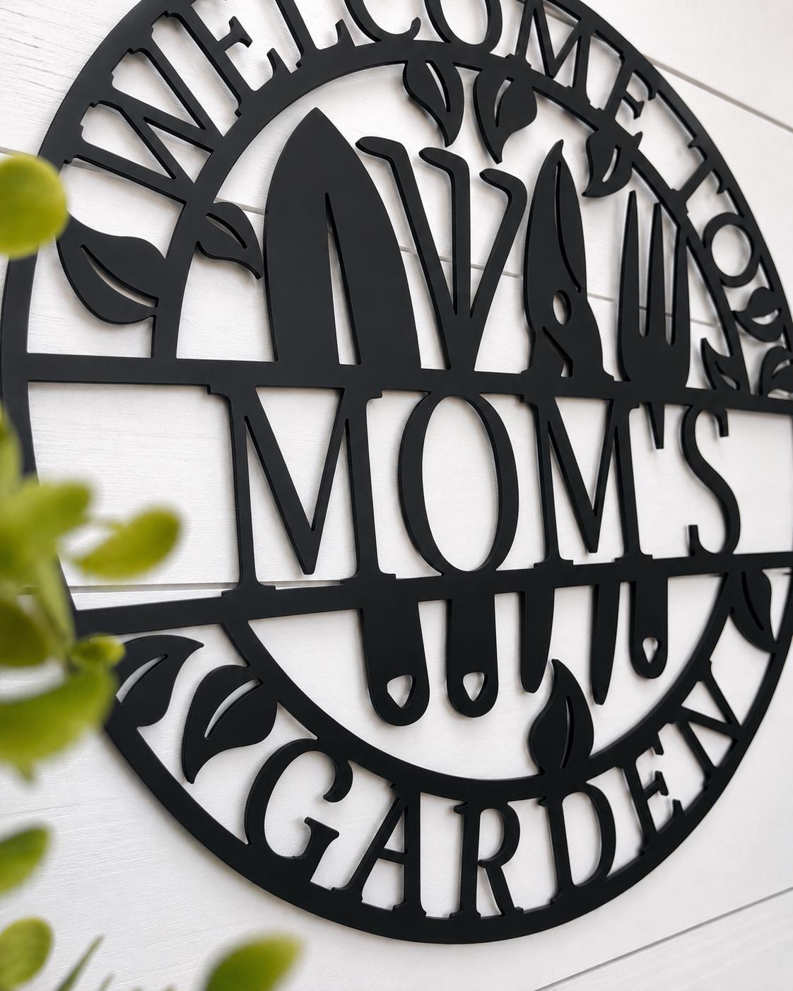 Black metal circular wall sign reading 'WELCOME TO MOM'S GARDEN' with gardening tool silhouettes, mounted on a white wood wall with blurred green leaves in the foreground.