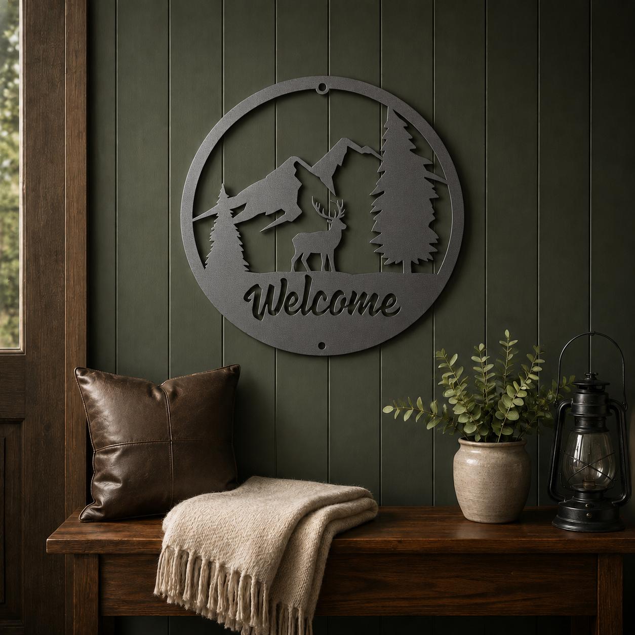 A circular metal wall sign with a mountain, tree, deer, and the word 'Welcome' cut out, hanging on a dark green paneled wall above a wooden bench with a brown pillow, beige throw blanket, and potted plant.