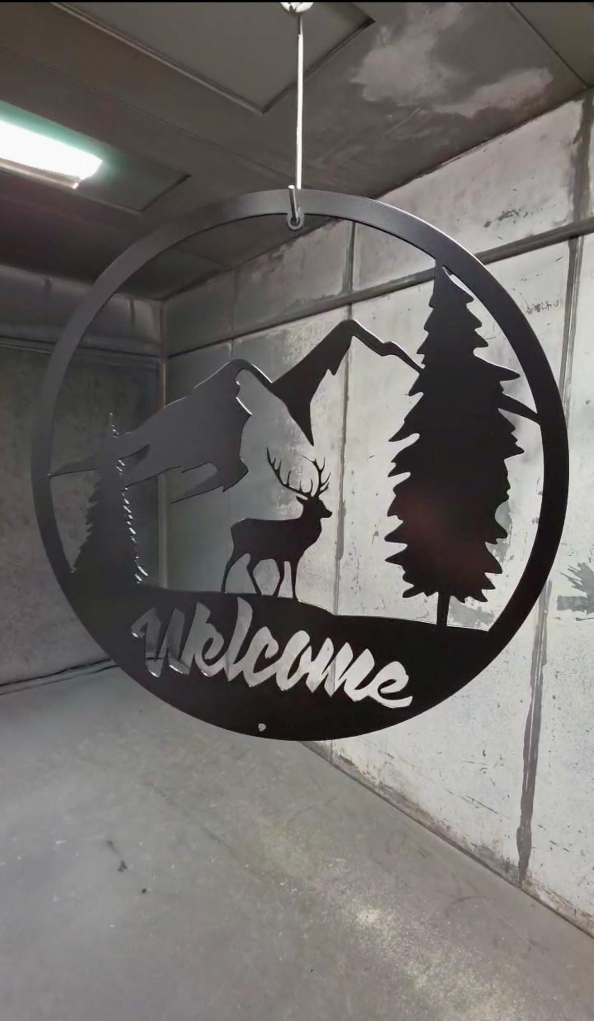 A circular black metal wall hanging sign with a silhouette of a deer, mountains, pine trees, and the word 'Welcome' in script, suspended by a wire in an indoor space.