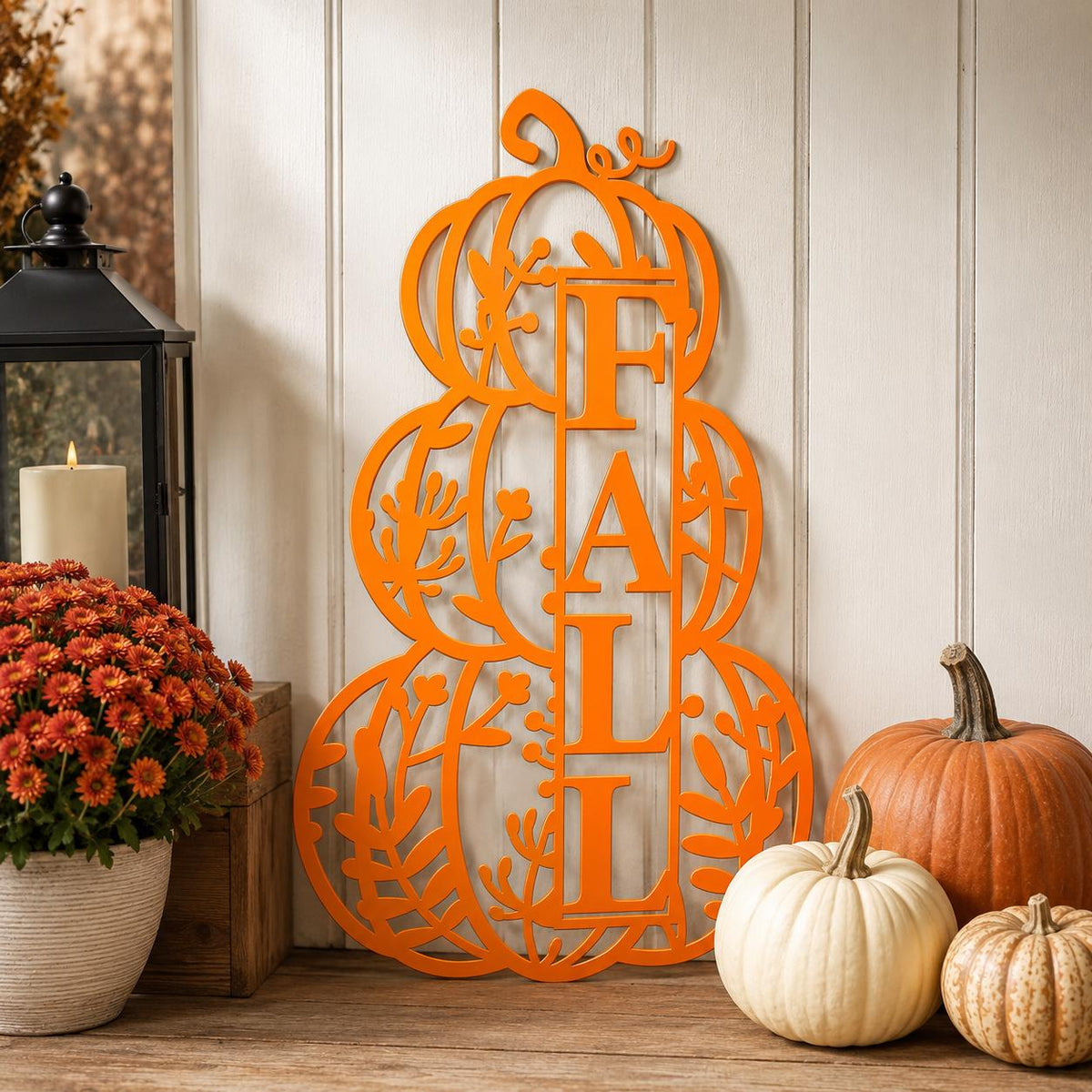 An orange metal decorative sign spelling FALL in stacked letters with leafy cutout details, displayed on a wooden surface against a white paneled wall, surrounded by pumpkins, a potted orange flower, and a black lantern.