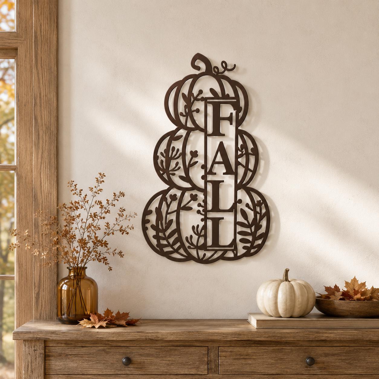 A dark brown metal wall decor piece shaped like stacked pumpkins with the word 'FALL' inside, displayed on a beige wall above a wooden console table with autumn decor.