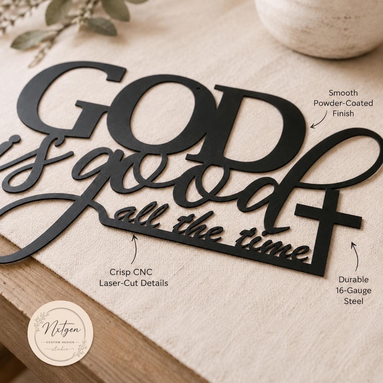 Black metal wall art sign reading “God is good all the time” with calligraphic lettering and a cross, shown on a beige fabric surface with decorative text callouts and a Nitzgen logo.