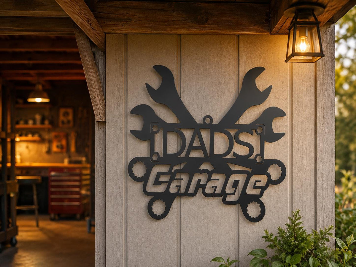 A black metal sign reading 'DAD'S Garage' with crossed wrenches behind the text, mounted on a light wood-paneled wall under a rustic wooden overhang. A warm lantern hangs to the right, and a blurred garage interior is visible in the background.