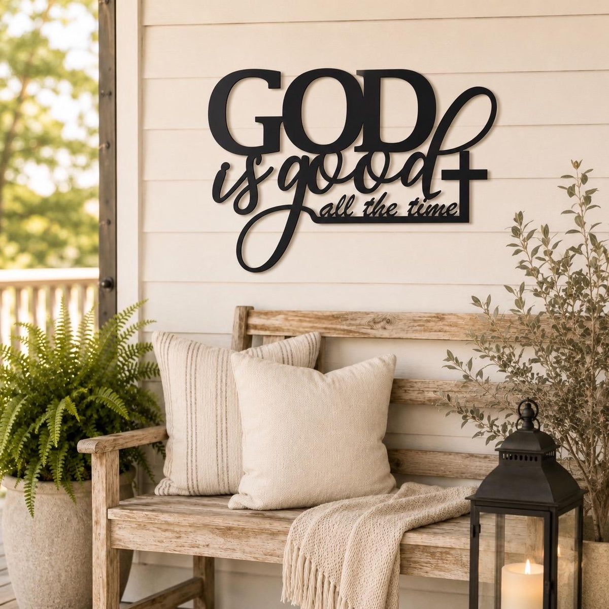 Black metal wall decor sign reading 'GOD is good all the time' mounted on a beige wood-paneled wall above a rustic wooden bench with cream pillows and a throw blanket, with a lantern and potted plants nearby.