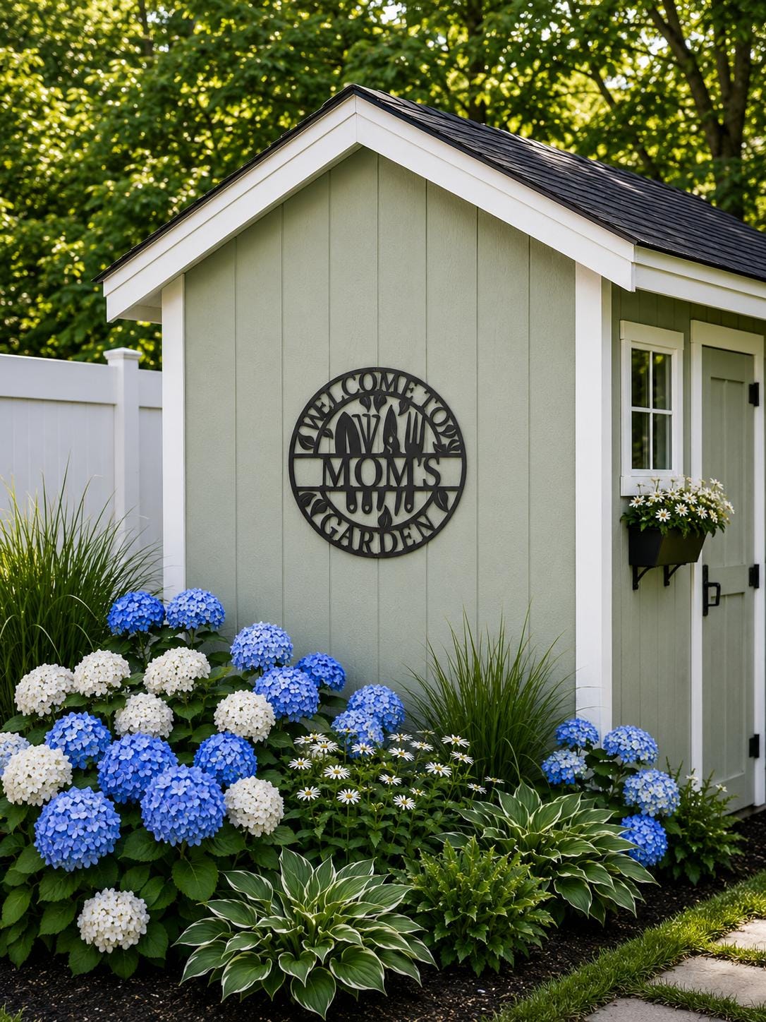 A small garden shed in a landscaped yard with blue and white hydrangeas in front. The shed is light sage green with white trim, a dark shingled roof, a white door, and a black metal sign reading 'Welcome to Mom's Garden'.